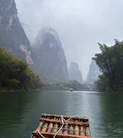 Balsa de bambú navegando por un río rodeado de montañas kársticas, vegetación exuberante y niebla, en un paisaje natural de China.