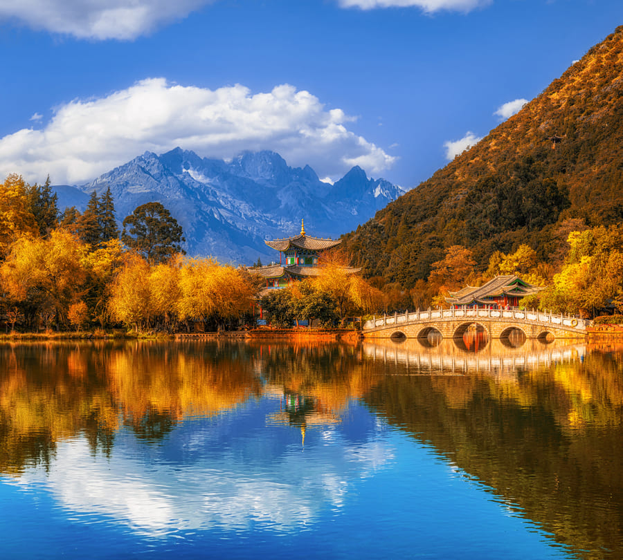 Lago con puente y pabellón tradicional chino rodeados de árboles otoñales y montañas al fondo.