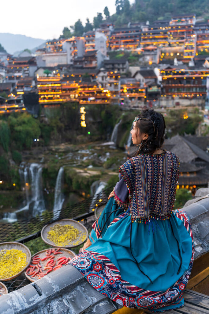 Persona con vestimenta tradicional observando un pueblo iluminado al atardecer, con cascadas y arquitectura típica sobre la ladera de una montaña.