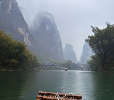 Balsa de bambú navegando por un río rodeado de montañas kársticas, vegetación exuberante y niebla, en un paisaje natural de China.